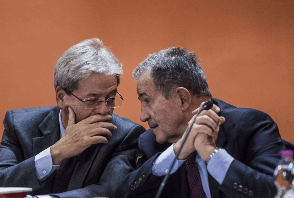 Paolo Gentiloni e Romano Prodi conversano durante un convegno politico, seduti al tavolo con sfondo arancione.