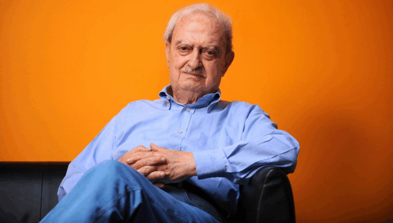 Emanuele Macaluso seduto su una poltrona, in primo piano, con sfondo arancione