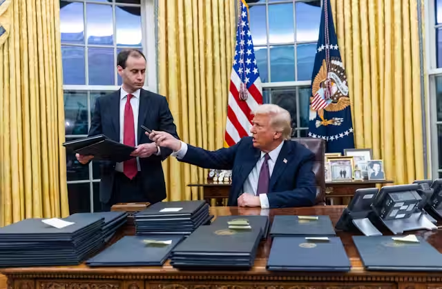 Donald Trump nell'Ufficio Ovale firma documenti ufficiali durante il suo secondo mandato presidenziale, circondato da faldoni e con un collaboratore.