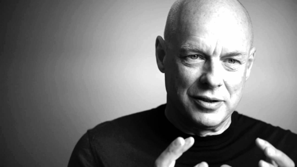 Brian Eno in un ritratto in bianco e nero, in primo piano mentre parla con gestualità espressiva.