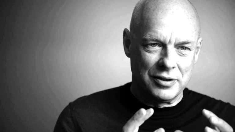 Brian Eno in un ritratto in bianco e nero, in primo piano mentre parla con gestualità espressiva.