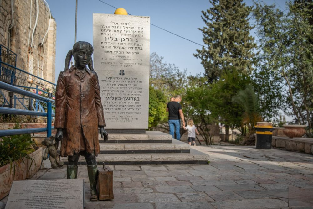 Statua commemorativa di una bambina con valigia davanti a un monumento dedicato alle vittime della Shoah, in un contesto urbano alberato.