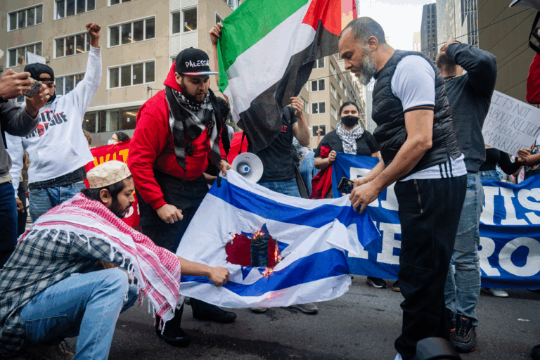 Manifestanti durante una protesta urbana calpestano una bandiera israeliana; sullo sfondo bandiere palestinesi e striscioni politici.