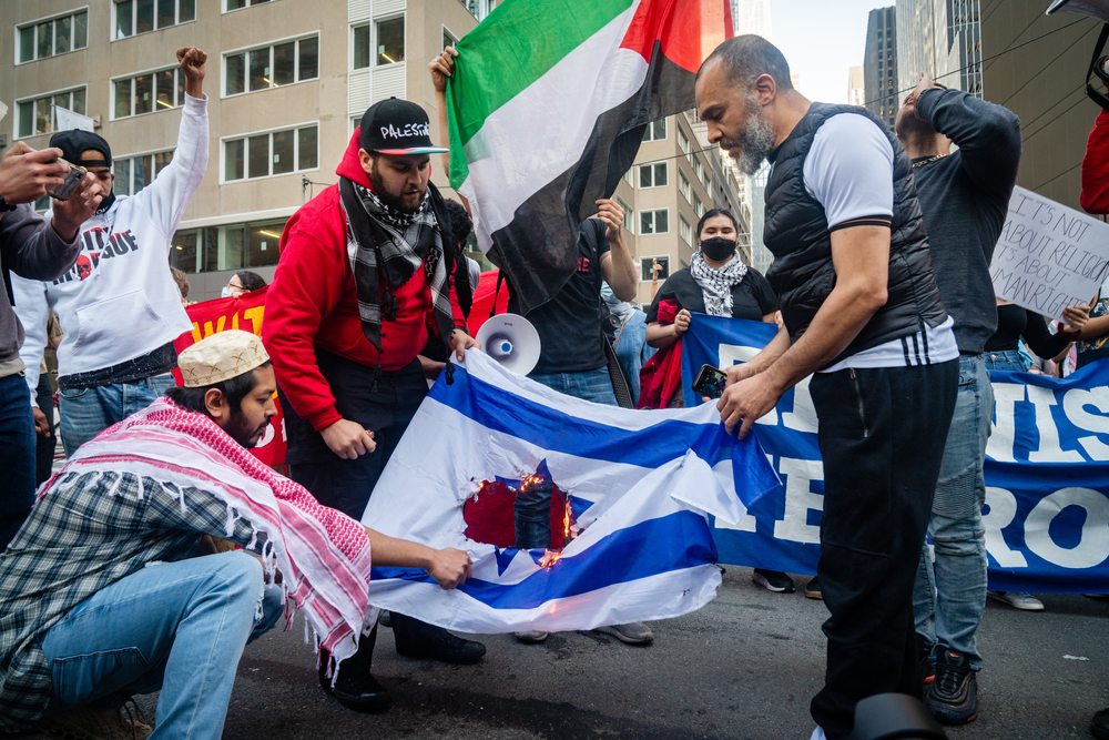 Manifestanti durante una protesta urbana calpestano una bandiera israeliana; sullo sfondo bandiere palestinesi e striscioni politici.
