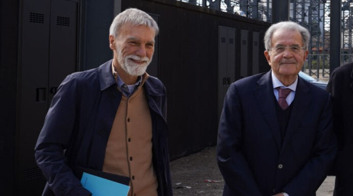 Due uomini anziani in abiti formali sorridono all’esterno durante un incontro pubblico o evento politico.