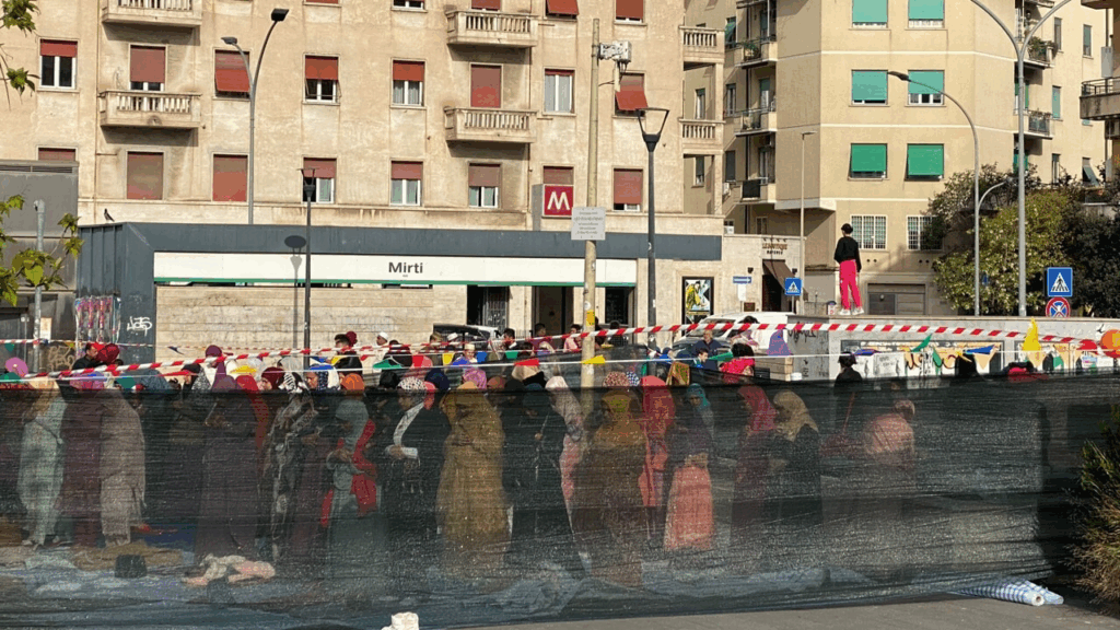 Donne migranti in fila davanti a un centro servizi in città, viste attraverso una rete di cantiere; sullo sfondo edifici popolari.
