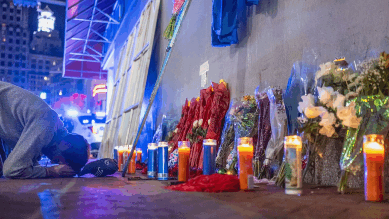 Uomo inginocchiato accanto a un memoriale con candele accese, fiori e luci blu, in ricordo delle vittime di un attacco.
