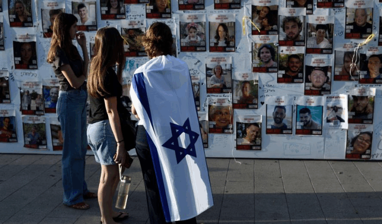Persone osservano un muro di foto degli ostaggi israeliani, una donna avvolta nella bandiera di Israele.