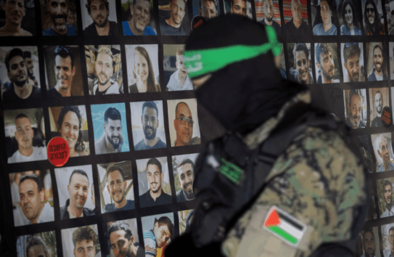 Uomo in uniforme militare con volto coperto davanti a una parete con fotografie di persone.