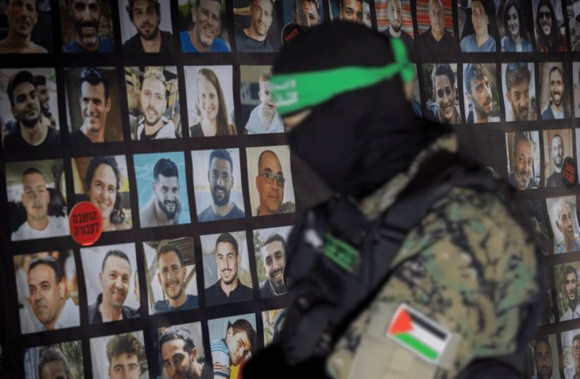 Uomo in uniforme militare con volto coperto davanti a una parete con fotografie di persone.