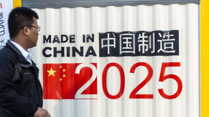 Uomo accanto a un cartellone con la scritta "Made in China 2025" e caratteri cinesi, simbolo della strategia industriale cinese.