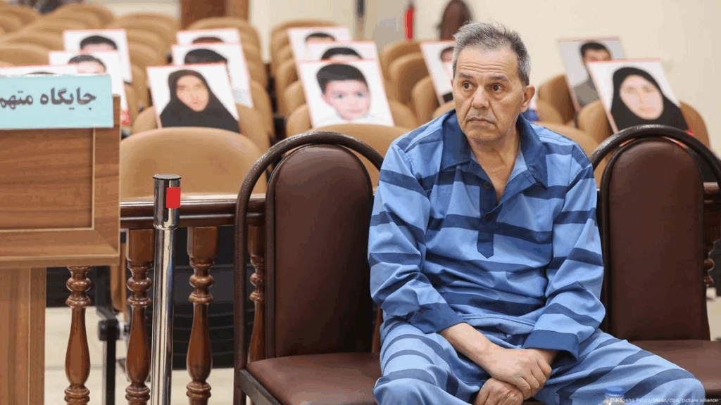 "Un uomo in abiti carcerari siede in aula di tribunale iraniana, mentre dietro di lui sono visibili sedie vuote con foto di persone, probabilmente vittime o oppositori."