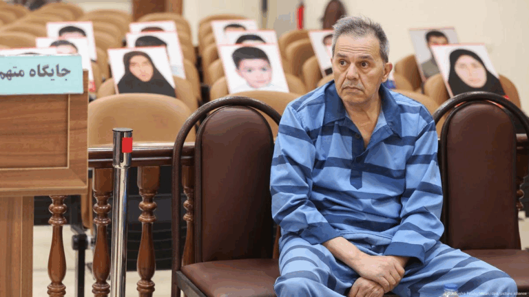 "Un uomo in abiti carcerari siede in aula di tribunale iraniana, mentre dietro di lui sono visibili sedie vuote con foto di persone, probabilmente vittime o oppositori."