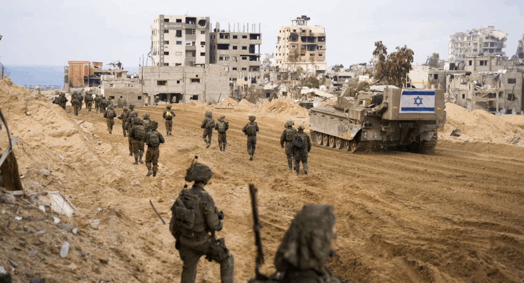 Soldati israeliani avanzano in una zona urbana distrutta durante un’operazione militare, con un carro armato e bandiera visibile.