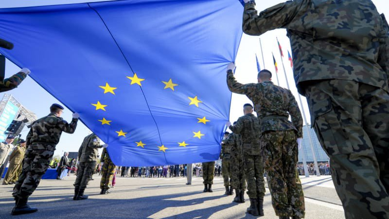 Soldati europei sollevano una grande bandiera dell’Unione Europea, simbolo dell’unità e della difesa comune nel contesto geopolitico globale.
