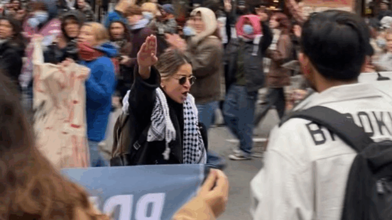 Persona al centro di una manifestazione affollata con il braccio alzato, circondata da cartelli e partecipanti, in un contesto urbano.