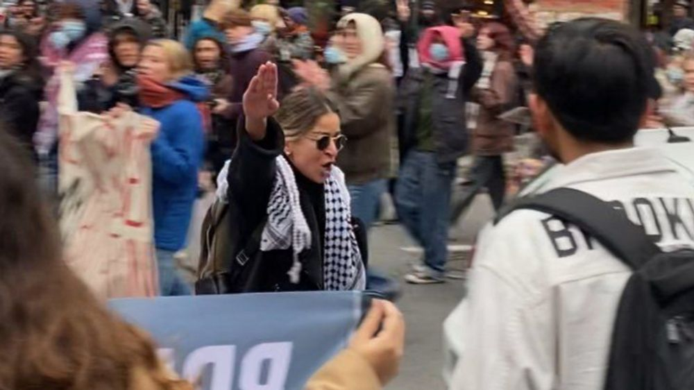 Persona al centro di una manifestazione affollata con il braccio alzato, circondata da cartelli e partecipanti, in un contesto urbano.