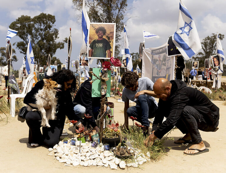 Persone inginocchiate depongono fiori su una tomba decorata con bandiere israeliane e la foto di un giovane, durante una commemorazione in Israele.
