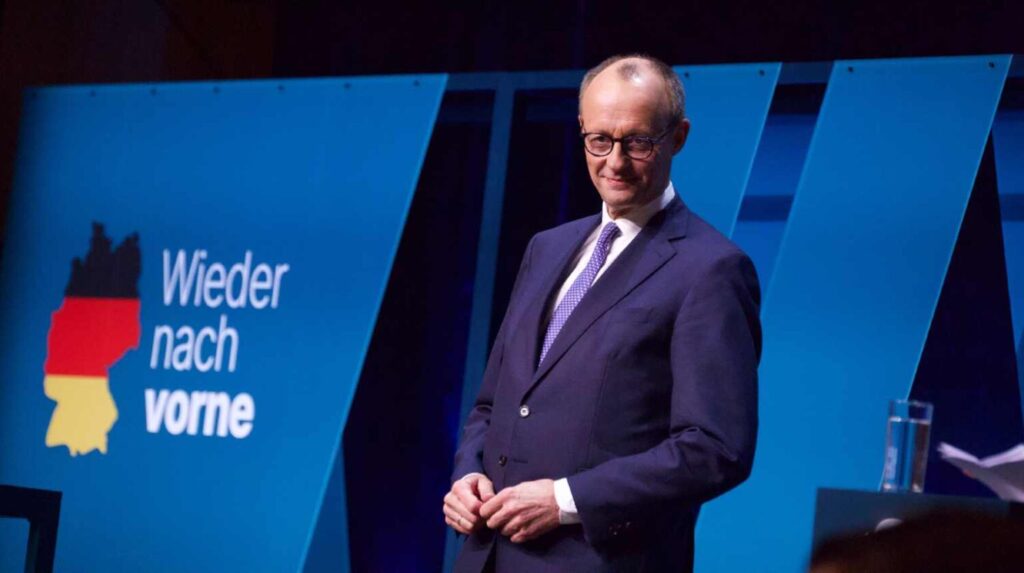 Friedrich Merz sul palco accanto a un pannello con la scritta “Wieder nach vorne” e una mappa della Germania stilizzata