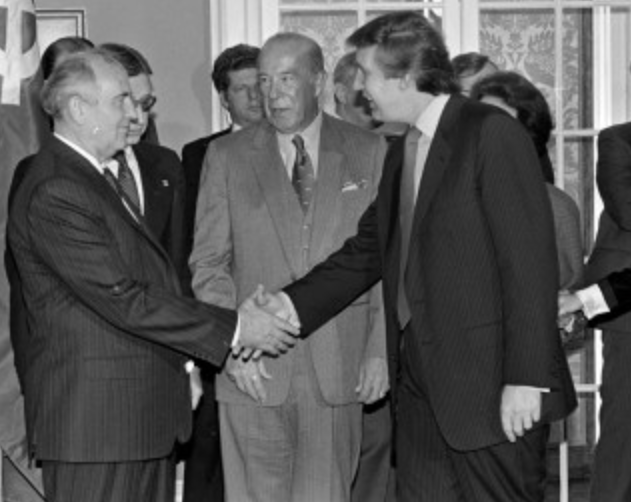 Donald Trump stringe la mano a un diplomatico sovietico durante un incontro ufficiale, circondato da altri uomini in abiti formali.