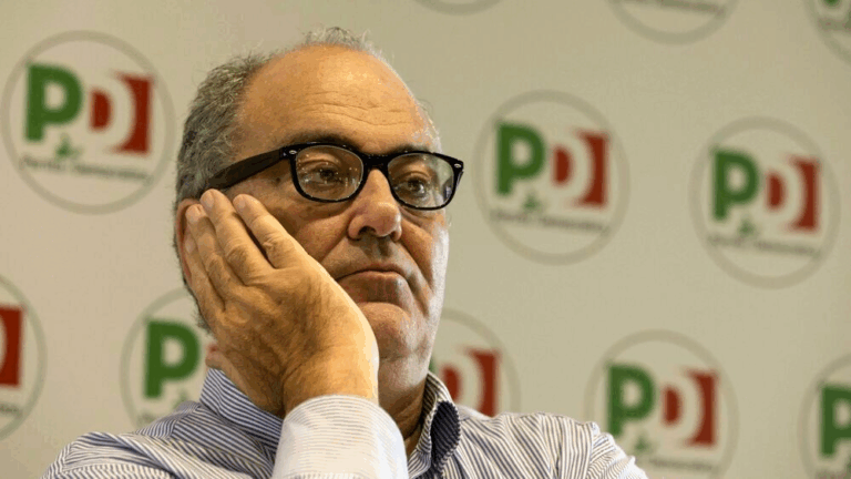 Goffredo Bettini con espressione riflessiva, davanti a uno sfondo con loghi del Partito Democratico.