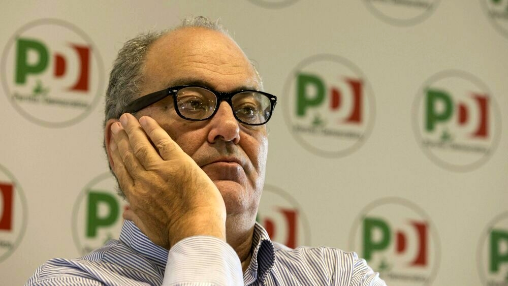 Goffredo Bettini con espressione riflessiva, davanti a uno sfondo con loghi del Partito Democratico.