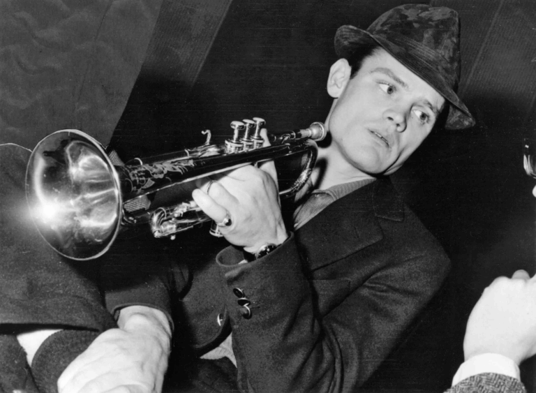 Chet Baker suona la tromba in una foto in bianco e nero, con cappello e abito elegante, immerso nell’atmosfera jazz del dopoguerra.