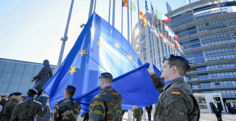 Militari europei con bandiera UE davanti al Parlamento: simbolo della sfida per una difesa comune e autonoma dell’Europa.