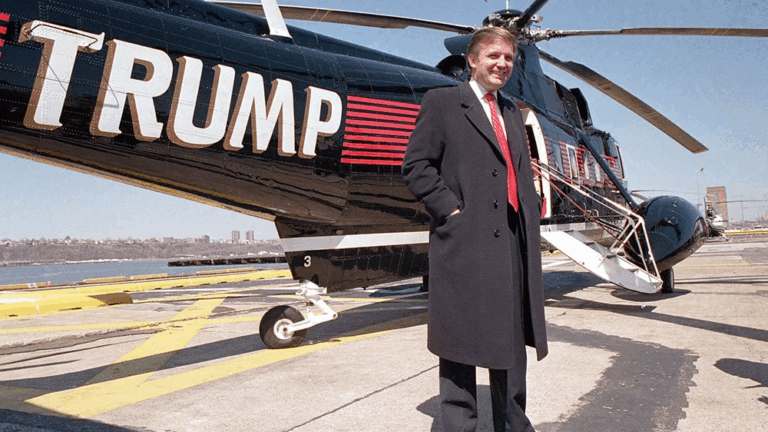 Donald Trump in piedi accanto a un elicottero nero con il suo nome scritto in grande in bianco e rosso, su una pista di atterraggio