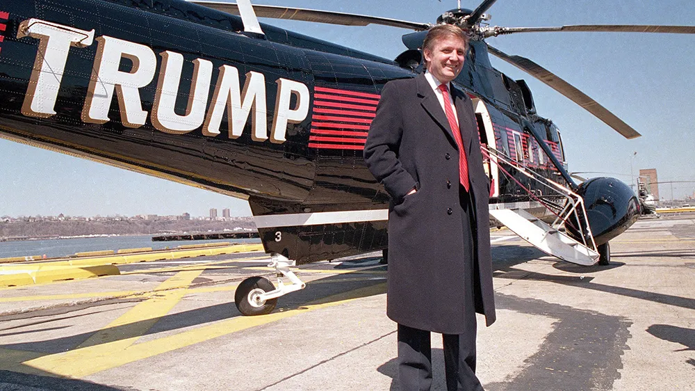 Donald Trump in piedi accanto a un elicottero nero con il suo nome scritto in grande in bianco e rosso, su una pista di atterraggio