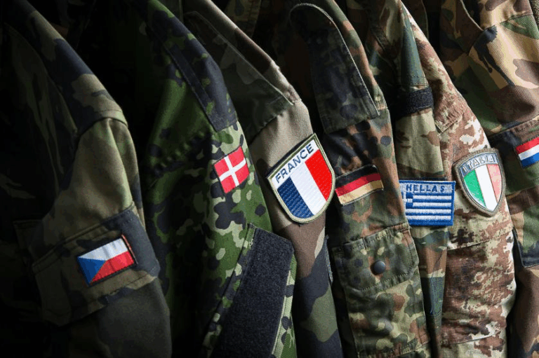 Uniformi militari di diversi paesi europei con bandiere nazionali cucite sulle maniche, simbolo della cooperazione nella difesa europea.