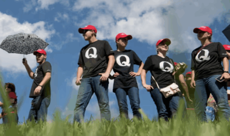 Persone con magliette QAnon e cappellini rossi marciano su un prato sotto un cielo azzurro, simbolo del movimento complottista americano.
