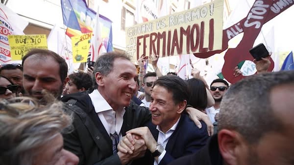 Giuseppe Conte tra la folla durante una manifestazione pubblica in Italia, con cartelli e slogan politici sullo sfondo