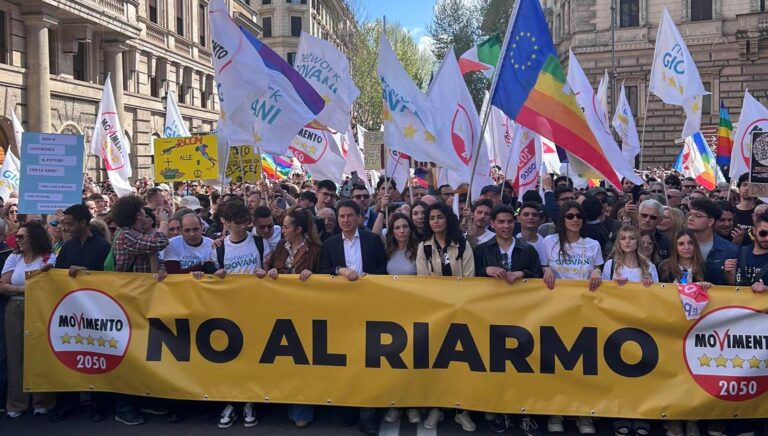Manifestazione pacifista con striscione "No al riarmo" e bandiere arcobaleno in una piazza italiana.